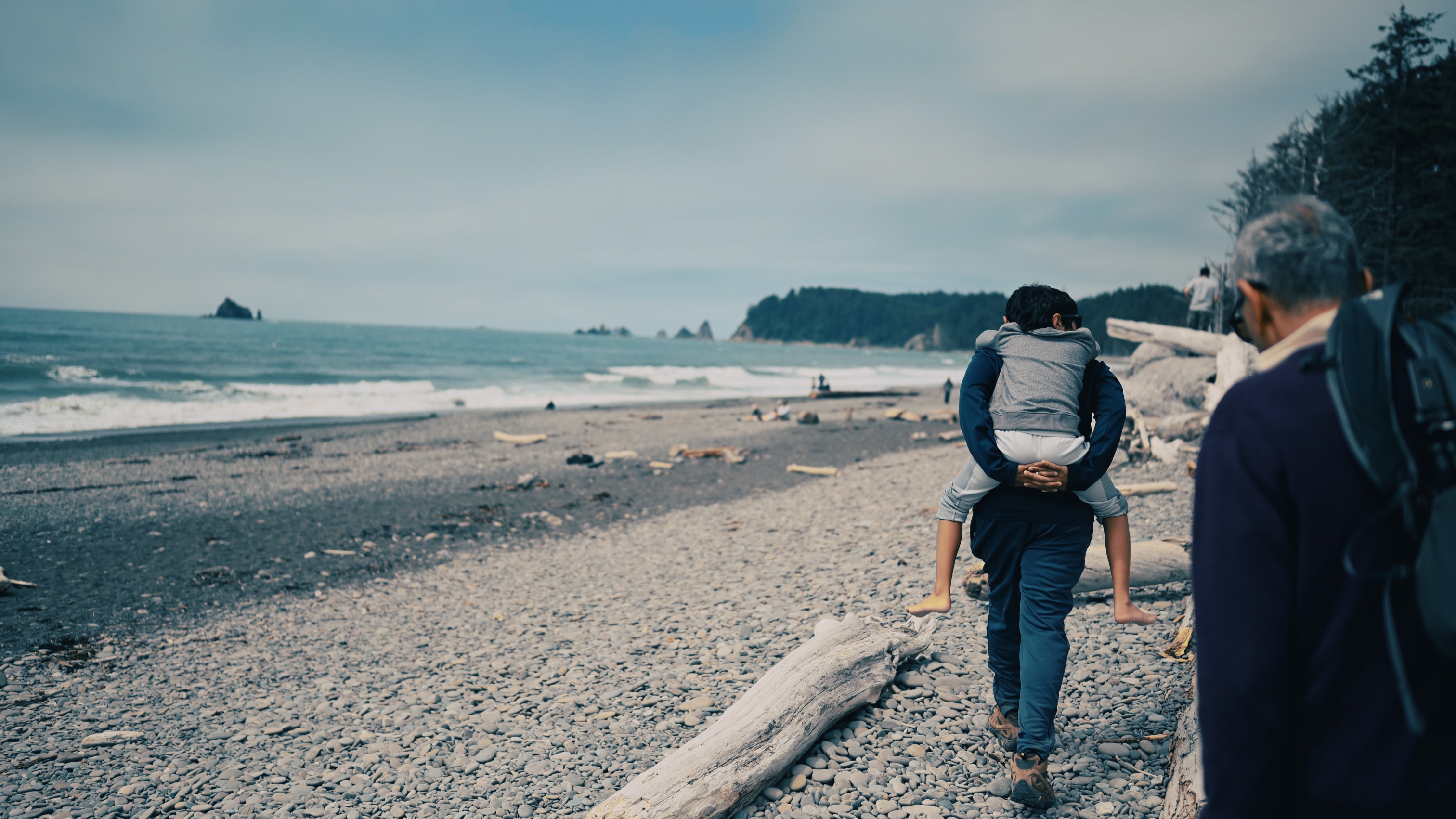 Man carrying a boy on his back walk along a pebbly beach with driftwood. Waves in the ocean, with sea cliffs and pine trees along the coastline. An elderly man is walking with a backpack.